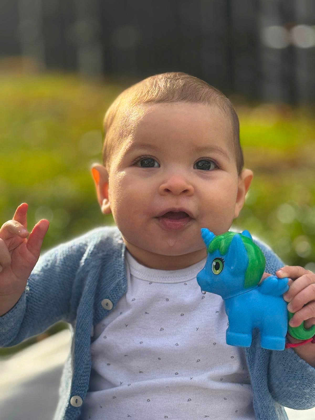 Laura a rejoint le concours — aidez-le/la à gagner de superbes lots ! baby, child, toy, unicorn, blue_cardigan, white_shirt, outdoor, portrait, cute, smiling, hand, face, young_child, nature, daylight, person, happy, clothing, infant, playful