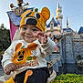 Riley joined the competition — help win amazing prizes! baby, child, infant, smiling, halloween, pumpkin_hat, pumpkin_sweatshirt, festive_clothing, castle, theme_park, anniversary_banner, outdoor, daylight, greenery, hand_reaching, person, cute, costume, happy, portrait