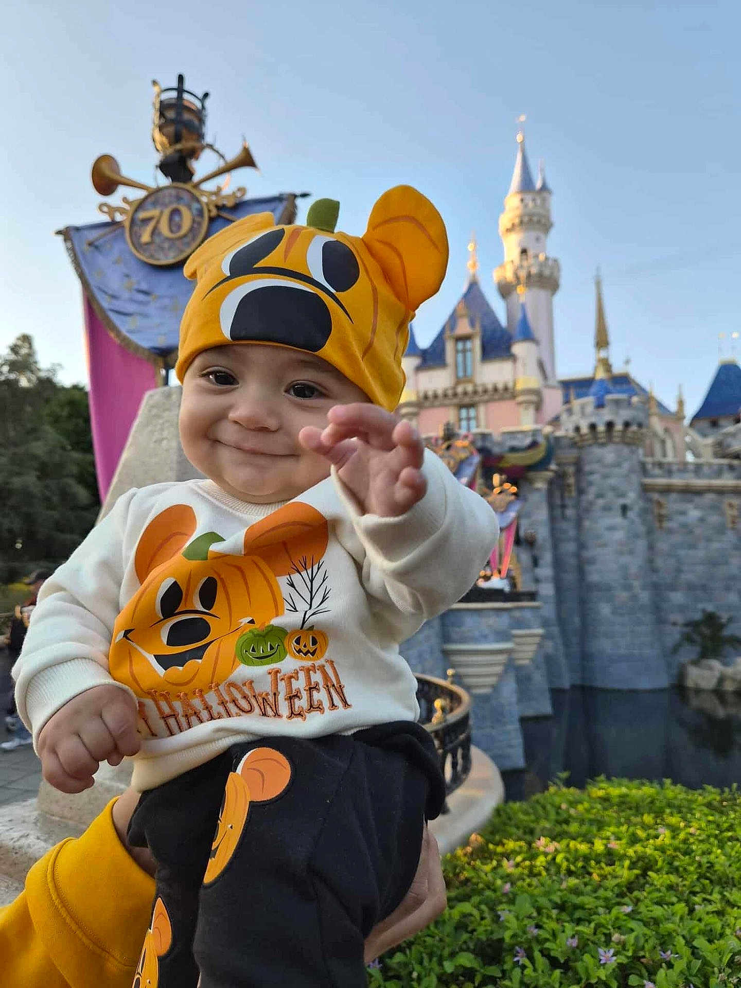 Riley joined the competition — help win amazing prizes! baby, child, infant, smiling, halloween, pumpkin_hat, pumpkin_sweatshirt, festive_clothing, castle, theme_park, anniversary_banner, outdoor, daylight, greenery, hand_reaching, person, cute, costume, happy, portrait