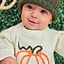 baby, child, knit_hat, sweater, pumpkin_design, smiling, face, portrait, closeup, hand, green_background, autumn, cute, cozy, warm_clothing, indoors, young_child, person, happy, headwear
