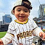 baby, child, smile, baseball, stadium, cap, jersey, sports, person, outdoor, fence, building, hand, clothing, happy, face, team, fan, infant, cute