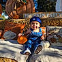 toddler, child, pumpkin, blue_cap, denim_overalls, smiling, hay_bale, fall_decor, pillows, outdoor, sunlight, autumn, cute, sitting, happy, seasonal, decorations, orange_pumpkin, white_pumpkin, casual_clothing