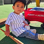 Riley joined the competition — help win amazing prizes! toddler, child, baseball, baseball_bat, cap, jeans, sneakers, toy_wagon, radio_flyer, outdoor, playground, grass, sunlight, person, sportswear, toy, sitting, young_child, recreation, casual_clothing