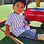 Riley is registered to the contest to win money with this photo: toddler, child, baseball, baseball_bat, cap, jeans, sneakers, toy_wagon, radio_flyer, outdoor, playground, grass, sunlight, person, sportswear, toy, sitting, young_child, recreation, casual_clothing