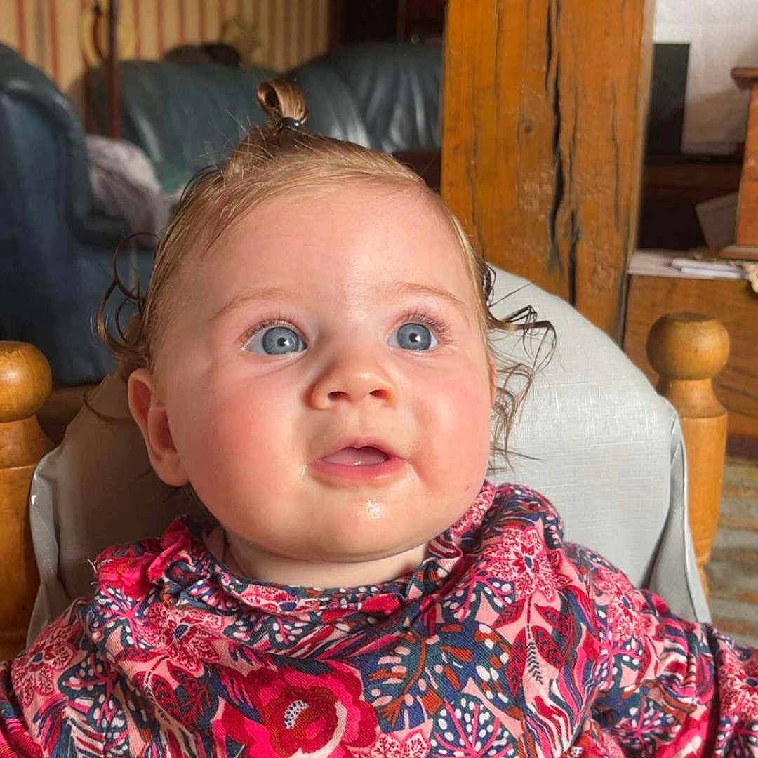 Laélya a rejoint le concours — aidez-le/la à gagner de superbes lots ! baby, blue_eyes, child, colorful_clothing, curious, curly_hair, cute, drool, expression, face, floral_dress, high_chair, indoor, infant, person, portrait, room, seated, topknot, wooden_furniture