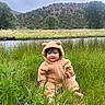 Presley joined the competition — help win amazing prizes! baby, child, grass, river, trees, hill, cloudy_sky, nature, outdoor, greenery, smiling, cute, onesie, fuzzy_clothing, portrait, sitting, peaceful, scenic, infant, cozy
