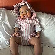 Presley joined the competition — help win amazing prizes! baby, infant, smile, pink, striped_clothing, bonnet, bed, white_bedding, red_wall, cute, chubby_legs, happy, child, portrait, indoors, person, sitting, soft_light, cozy, young_child