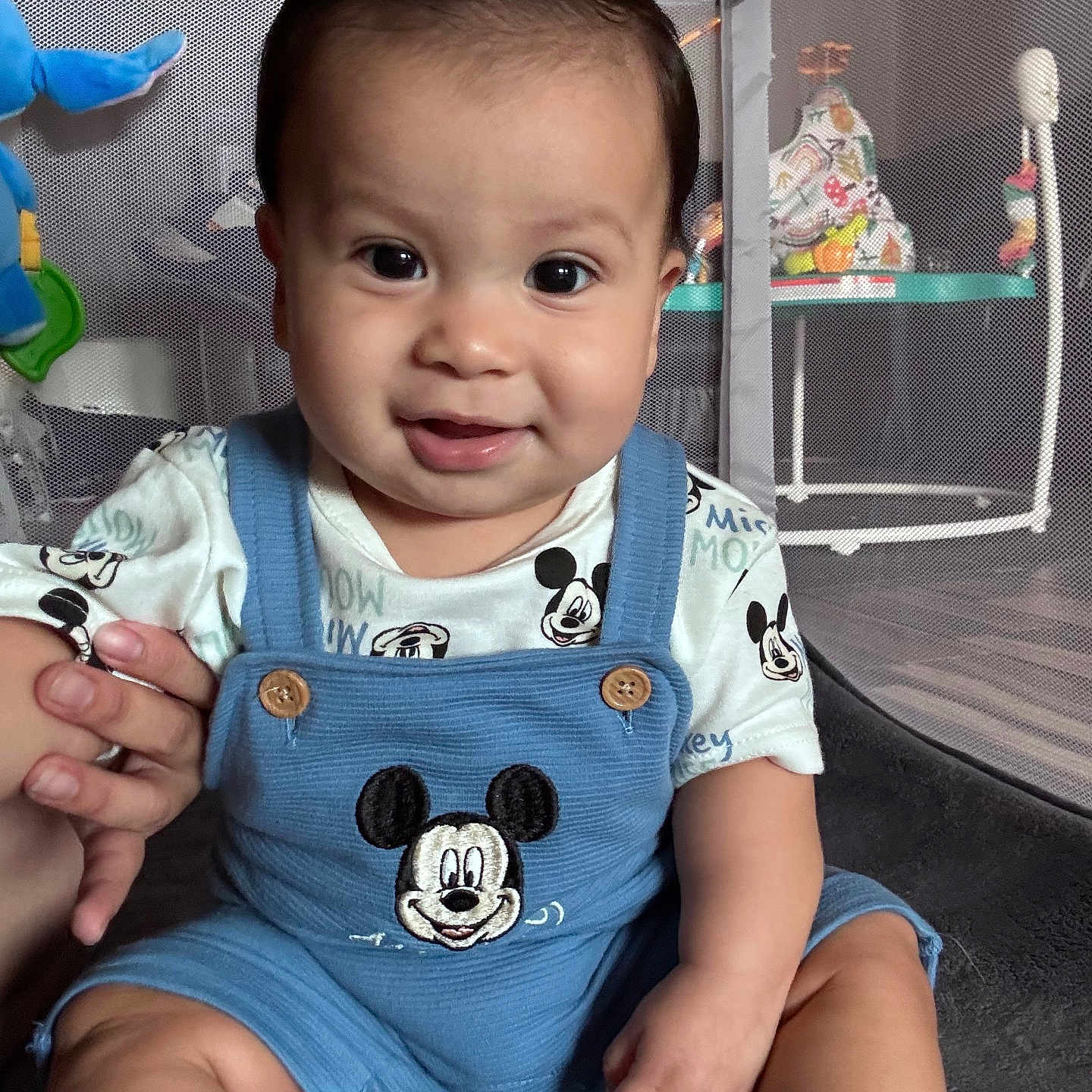 Lukas is registered to the contest to win money with this photo: baby, child, infant, mickey_mouse, overalls, blue_clothing, smiling, holding_hand, playpen, toy, indoor, cute, happy, person, face, sitting, short_hair, closeup, skin, young