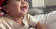 Lukas is registered to the contest to win money with this photo: baby, child, smiling, red_cap, bedroom, crib, pillow, mattress, quilted, infant, cute, happy, indoor, person, clothing, hand, face, tongue_out, headwear, relaxing