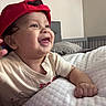 Lukas is registered to the contest to win money with this photo: baby, child, smiling, red_cap, bedroom, crib, pillow, mattress, quilted, infant, cute, happy, indoor, person, clothing, hand, face, tongue_out, headwear, relaxing