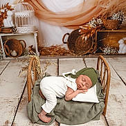 Maé participe au concours pour gagner de l'argent avec cette photo : baby, sleeping, bed, blanket, pillow, wooden_floor, woven_basket, dried_flowers, green_cap, white_outfit, nursery, rustic, decor, soft_lighting, peaceful, infant, portrait, cozy, fabric, wood