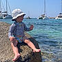 Maé a rejoint le concours — aidez-le/la à gagner de superbes lots ! toddler, child, rock, water, sea, sun_hat, striped_shirt, boats, sailboats, clear_sky, sunny, outdoor, coast, shore, summer, vacation, happy, barefoot, nature, daytime