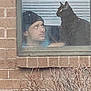 person, cat, window, brick_wall, beanie, indoor, pet, looking, black_cat, face, animal, glass, bush, plant, daytime, house, window_blinds, sitting, headwear, reflection