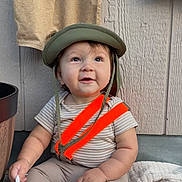Joaquin is registered to the contest to win money with this photo: baby, blanket, child, clothing, curious_expression, cute, face, happy, helmet, holding_object, indoor, infant, orange_suspender, pants, person, sitting, smiling, soft_light, striped_shirt, wooden_wall