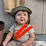 baby, blanket, child, clothing, curious_expression, cute, face, happy, helmet, holding_object, indoor, infant, orange_suspender, pants, person, sitting, smiling, soft_light, striped_shirt, wooden_wall