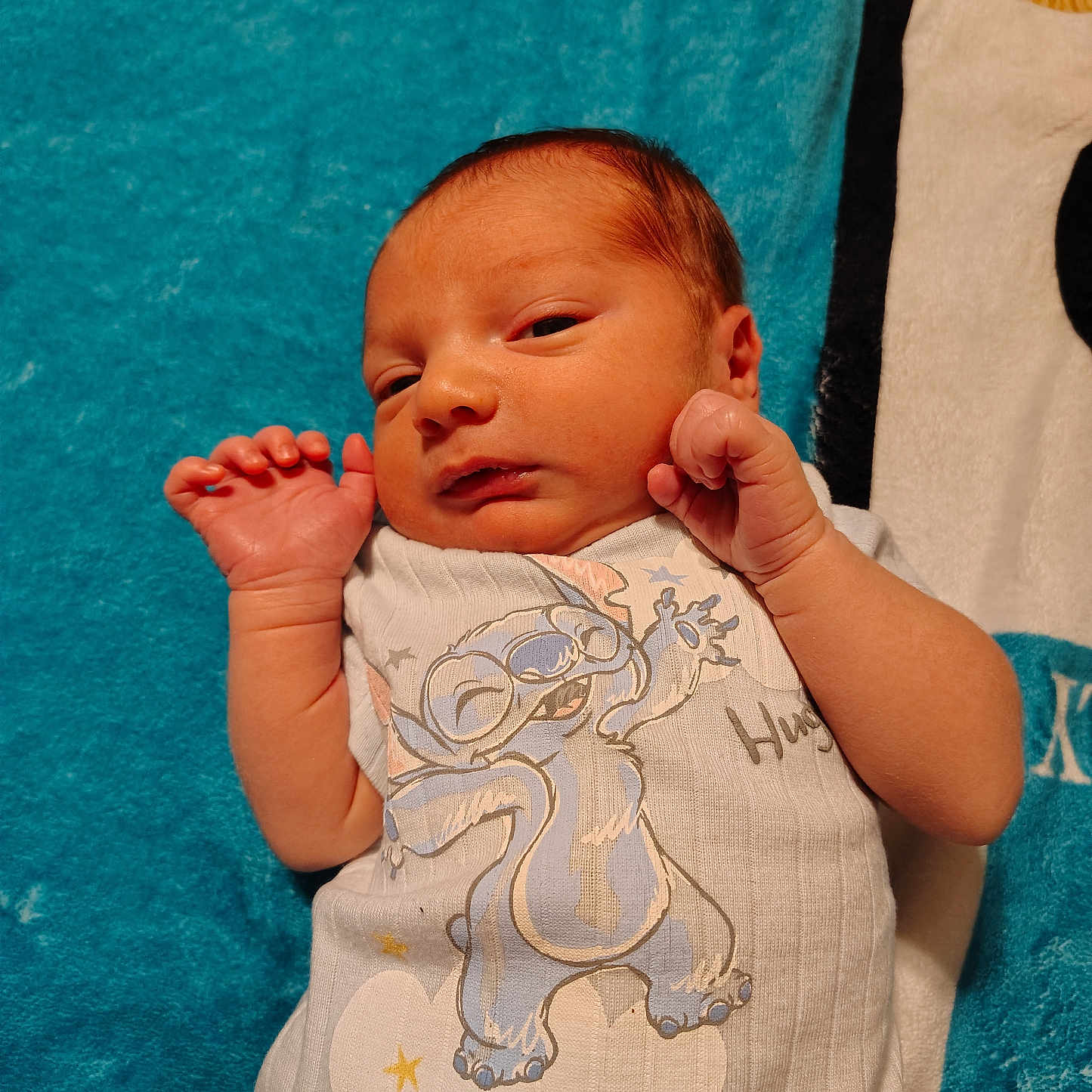 Luther is registered to the contest to win money with this photo: baby, blanket, blue, cartoon, child, clothing, cute, expression, face, hand, infant, lying_down, newborn, onesie, person, portrait, skin, sleepy, white, wood_floor