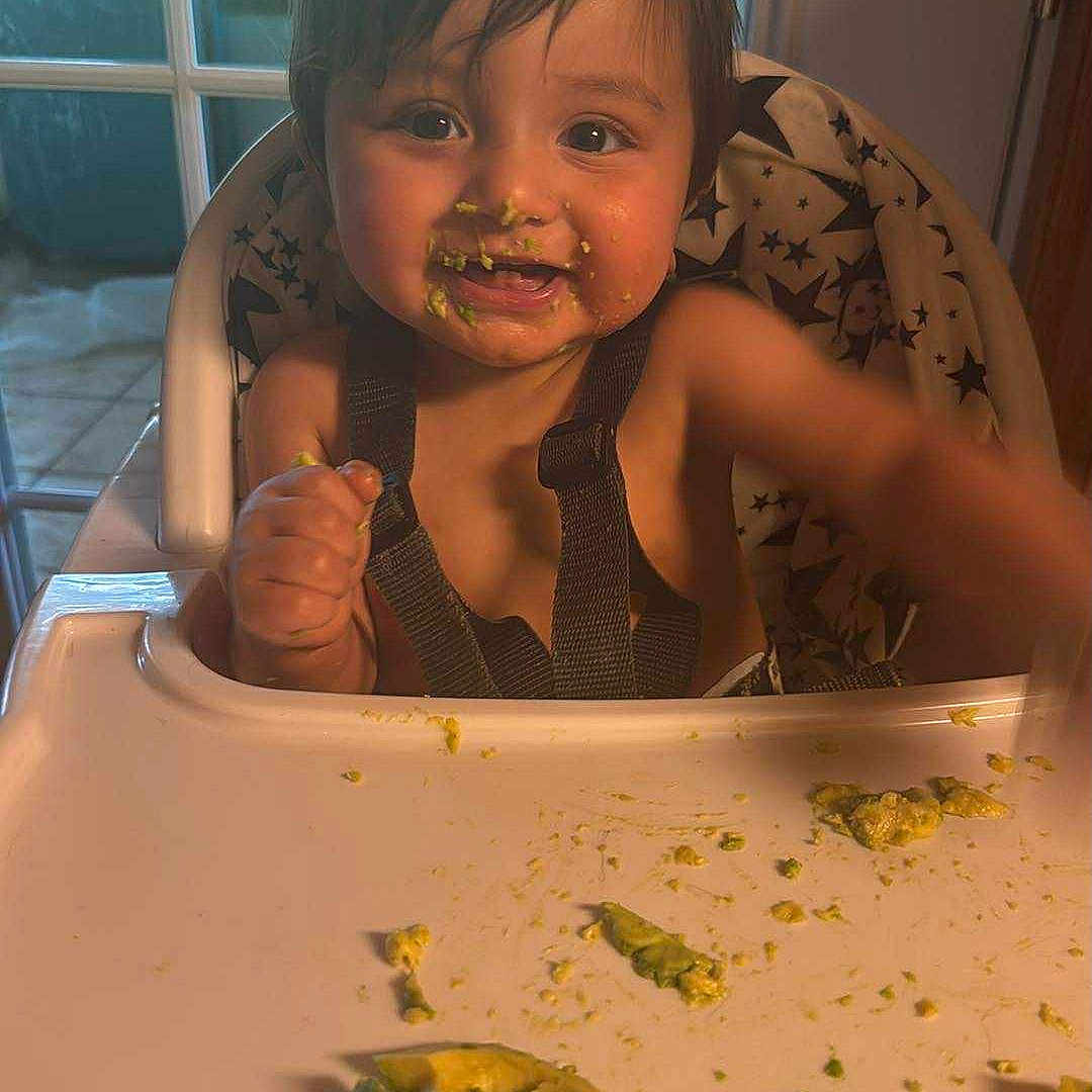 Ysmael is registered to the contest to win money with this photo: avocado, baby, child, cushion, cute, eating, face, food, hair, hand, happy, high_chair, indoor, infant, messy_face, smiling, star_pattern, table, tray, window