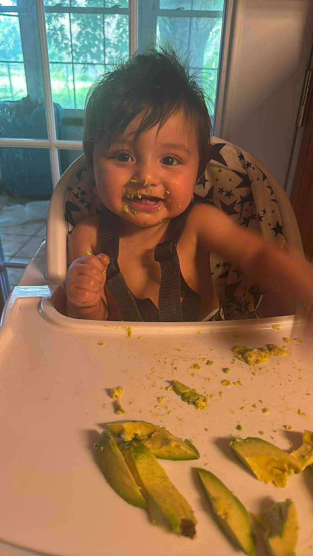 Ysmael is registered to the contest to win money with this photo: baby, high_chair, food, avocado, messy_face, child, indoor, window, star_pattern, cushion, tray, happy, cute, eating, smiling, hair, face, hand, table, infant