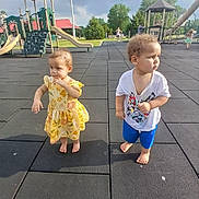 Kelsey is registered to the contest to win money with this photo: child, toddler, playground, outdoor, barefoot, yellow_dress, blue_shorts, cartoon_tshirt, slide, climbing_structure, rubber_mat, summer, park, curly_hair, grass, trees, play_equipment, person, daylight, casual_clothing