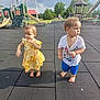 child, toddler, playground, outdoor, barefoot, yellow_dress, blue_shorts, cartoon_tshirt, slide, climbing_structure, rubber_mat, summer, park, curly_hair, grass, trees, play_equipment, person, daylight, casual_clothing