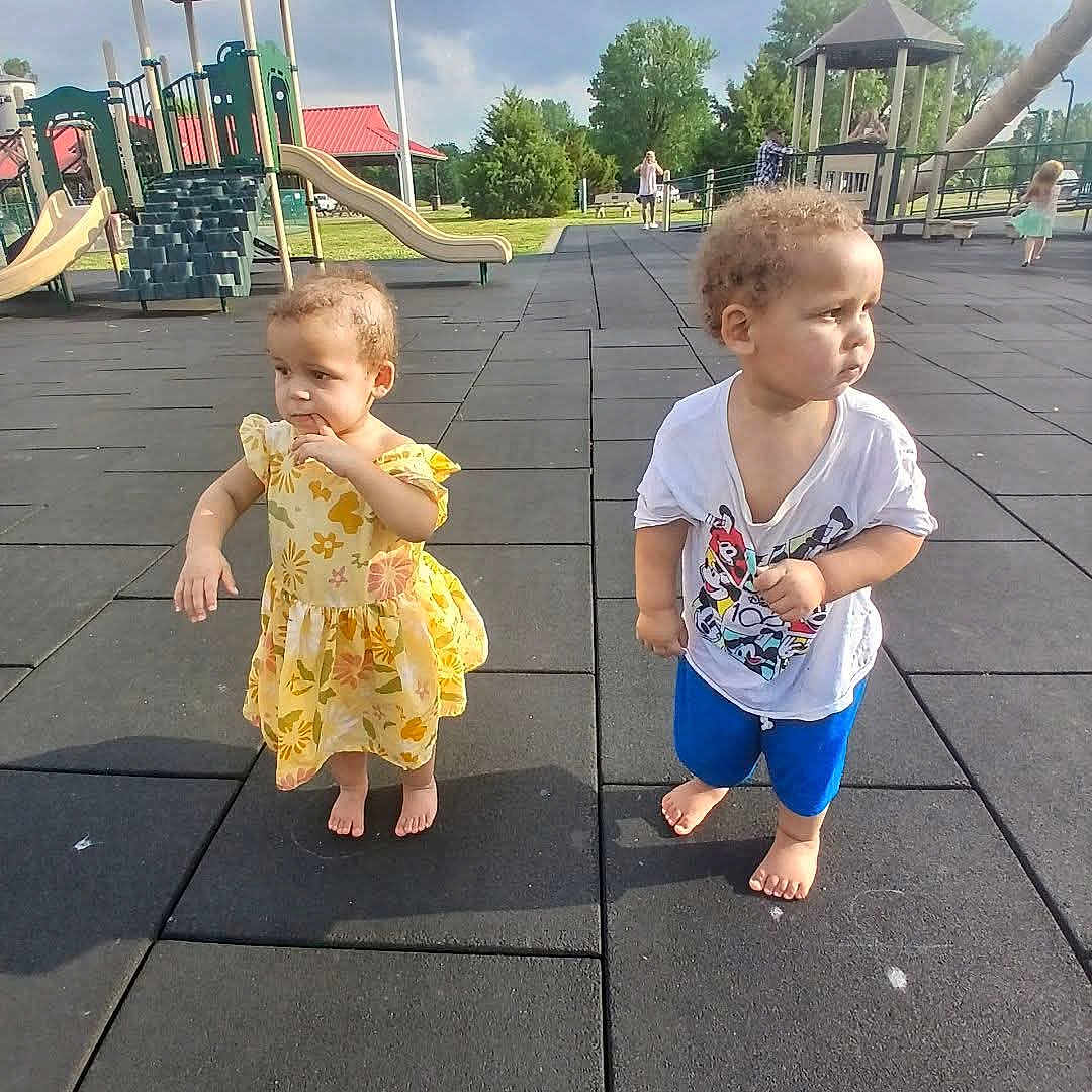 Kelsey is registered to the contest to win money with this photo: child, toddler, playground, outdoor, barefoot, yellow_dress, blue_shorts, cartoon_tshirt, slide, climbing_structure, rubber_mat, summer, park, curly_hair, grass, trees, play_equipment, person, daylight, casual_clothing