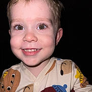 Adil participe au concours pour gagner de l'argent avec cette photo : child, toddler, face, smile, eyes, blonde_hair, pajamas, close_up, portrait, teeth, nose, cheeks, flash, indoor, dark_background, happy, cute, curious, patterned_clothing, button