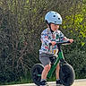 child, helmet, balance_bike, outdoor, greenery, pavement, sunlight, shorts, sneakers, trees, nature, happy, play, fun, summer, boy, casual_clothing, daytime, exercise, park