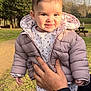 baby, child, clothing, cute, face, floral_pattern, grass, hand, happy, jacket, nature, outdoor, park, person, picnic_table, puffy_jacket, smiling, sunlight, trees, winter_clothes