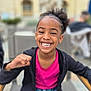 child, girl, smiling, happy, outdoor, portrait, curly_hair, jacket, pink_shirt, chair, casual, daylight, urban, background_blur, person, seated, young, gesture, joyful, candid