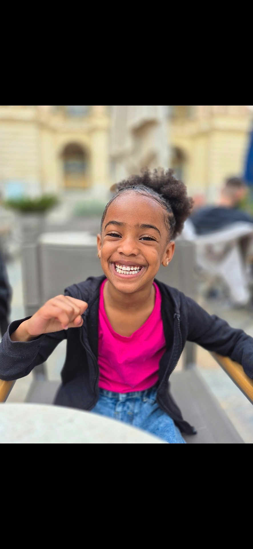 Emila participe au concours pour gagner de l'argent avec cette photo : child, girl, smiling, happy, outdoor, portrait, curly_hair, jacket, pink_shirt, chair, casual, daylight, urban, background_blur, person, seated, young, gesture, joyful, candid