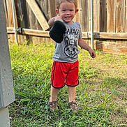 Elian is registered to the contest to win money with this photo: child, boy, smile, shoe, grass, fence, outdoor, tshirt, shorts, footwear, happy, person, summer, playful, young, cute, nature, casual_clothing, daylight, portrait