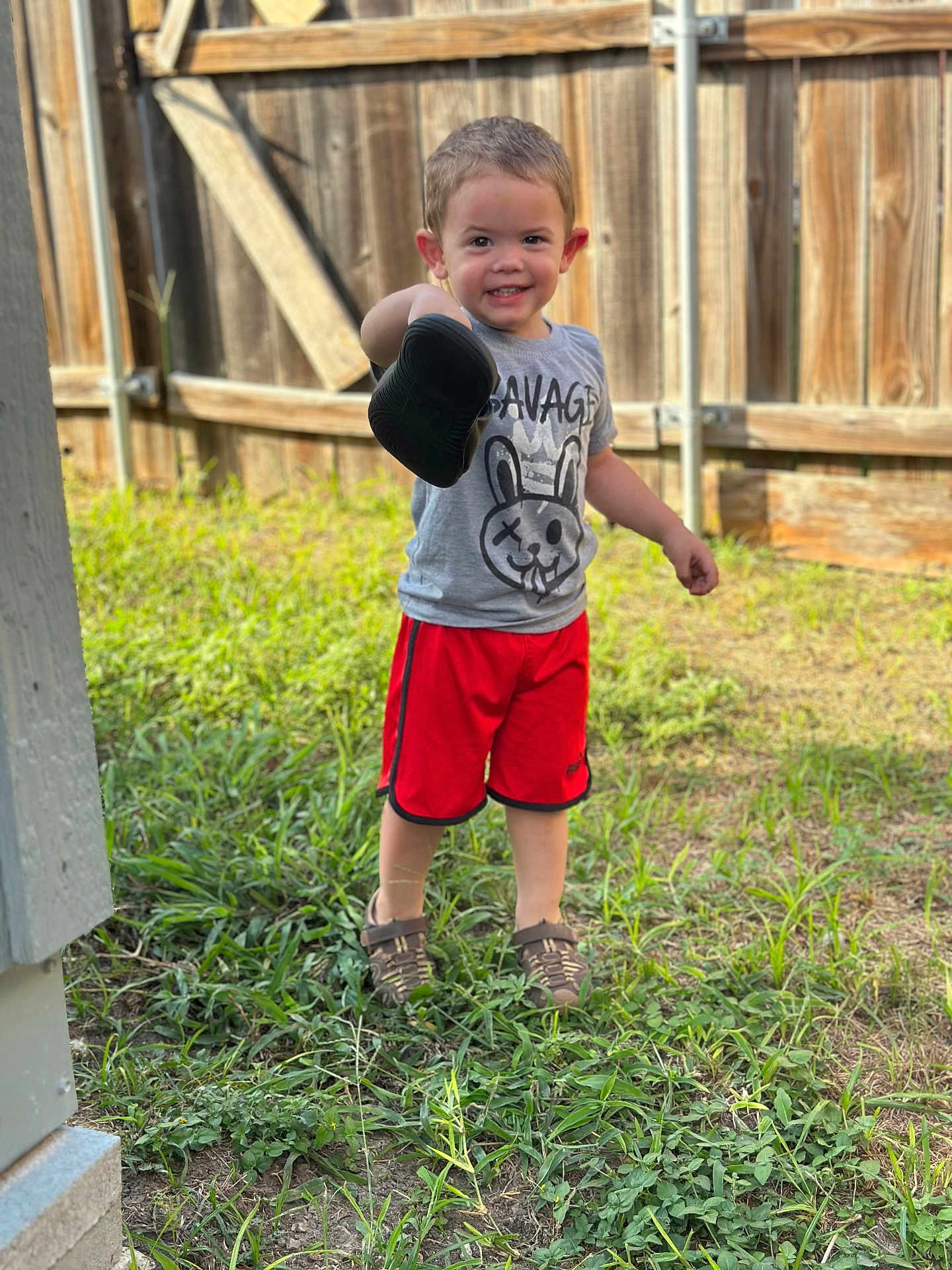 Elian is registered to the contest to win money with this photo: child, boy, smile, shoe, grass, fence, outdoor, tshirt, shorts, footwear, happy, person, summer, playful, young, cute, nature, casual_clothing, daylight, portrait