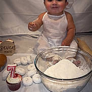 Mario joined the competition — help win amazing prizes! baby, flour, baking, cupcake, donuts, rolling_pin, white_clothing, baby_hat, tablecloth, celebration, food, powdered_sugar, smiling, portrait, sitting, kitchen, child, birthday, seven_months, cute