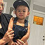 toddler, child, face, cap, denim_overalls, pout, adult_hand, indoor, door, window, wall, photograph_frame, person, expression, holding, nail_art, bracelet, skin, clothing, portrait