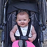 baby, stroller, child, smiling, pink_pants, white_top, seatbelt, outdoor, happy, infant, face, cute, person, portrait, baby_clothing, sitting, baby_girl, sunlight, daytime, playful