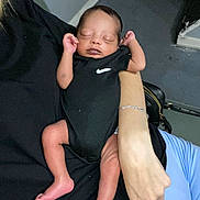 Kyrie is registered to the contest to win money with this photo: baby, newborn, sleeping, person, arm, bracelet, clothing, black_onesie, nike_logo, infant, skin, hand, foot, indoor, floor, bag, holding, resting, peaceful, casual