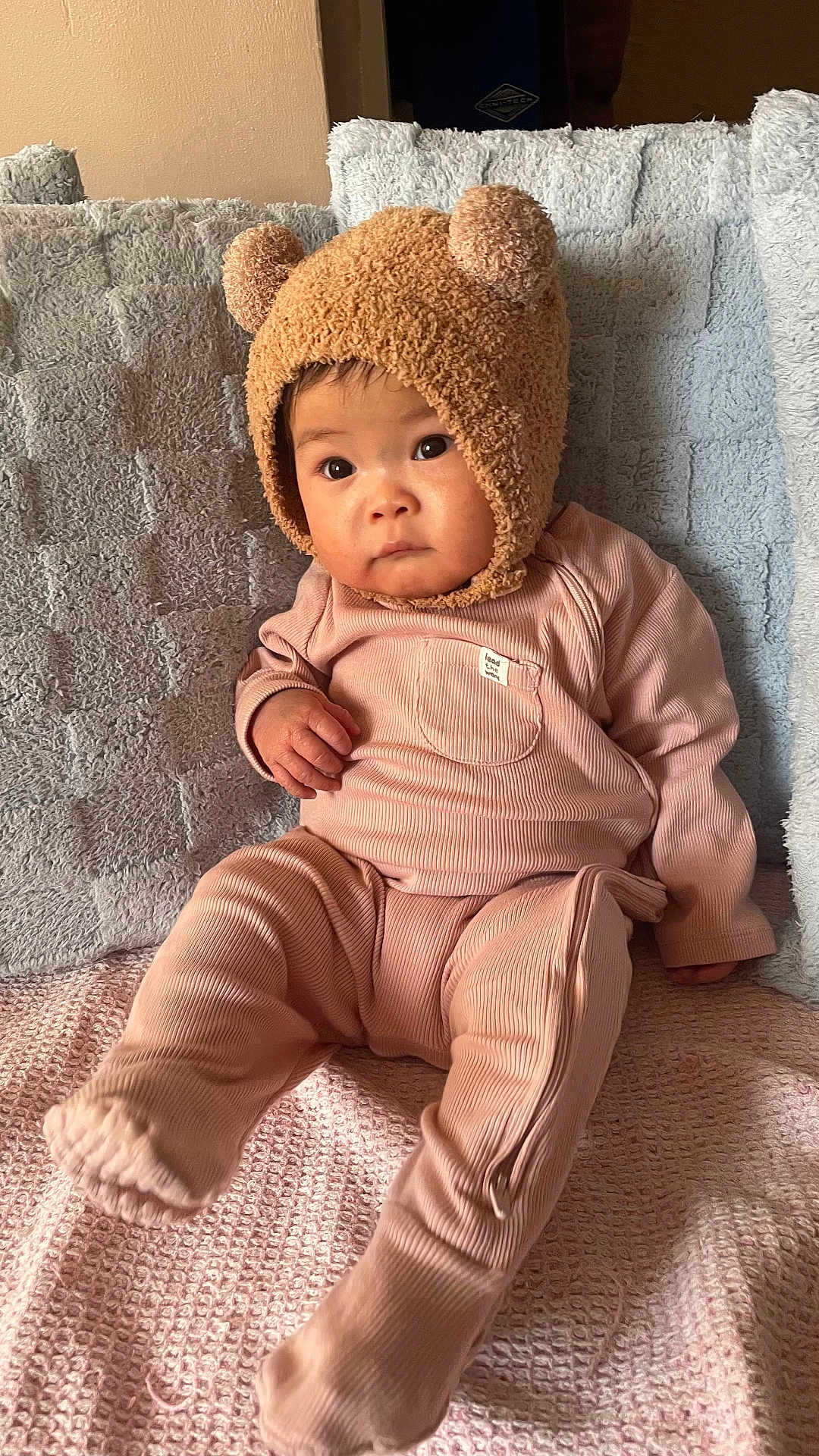 Eloise joined the competition — help win amazing prizes! baby, child, infant, pink_outfit, bear_hat, hat_with_ears, cute, indoors, couch, blanket, textured_blanket, seated, portrait, looking_away, soft_lighting, warm_colors, clothing, fuzzy_hat, expression, young_child