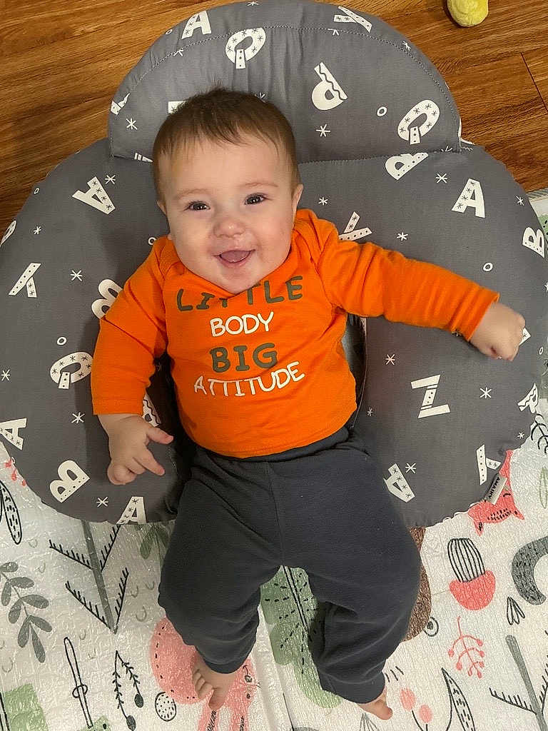 Jameson joined the competition — help win amazing prizes! baby, smiling, orange_shirt, cushion, alphabet_prints, play_mat, wooden_floor, happy, child, infant, clothing, flooring, pattern, person, cute, indoors, toddler, laying_down, text_on_clothing, pillow