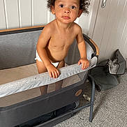 Remii joined the competition — help win amazing prizes! toddler, child, curly_hair, playpen, diaper, indoor, carpet, white_wall, curious, standing, looking_up, bare_chest, furniture, room, handrail, expression, young_child, home, door, gray
