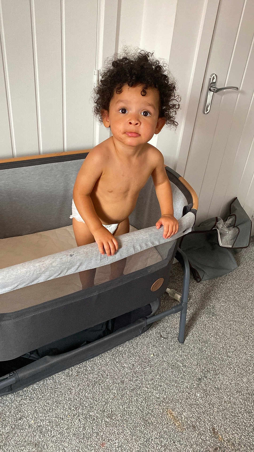 Remii joined the competition — help win amazing prizes! toddler, child, curly_hair, playpen, diaper, indoor, carpet, white_wall, curious, standing, looking_up, bare_chest, furniture, room, handrail, expression, young_child, home, door, gray