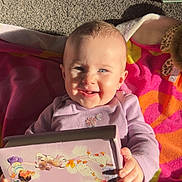 Josie is registered to the contest to win money with this photo: baby, child, smiling, book, reading, blanket, carpet, indoors, sunlight, happy, portrait, infant, clothing, hand, face, eyewear, toys, soft_toys, colorful, cute