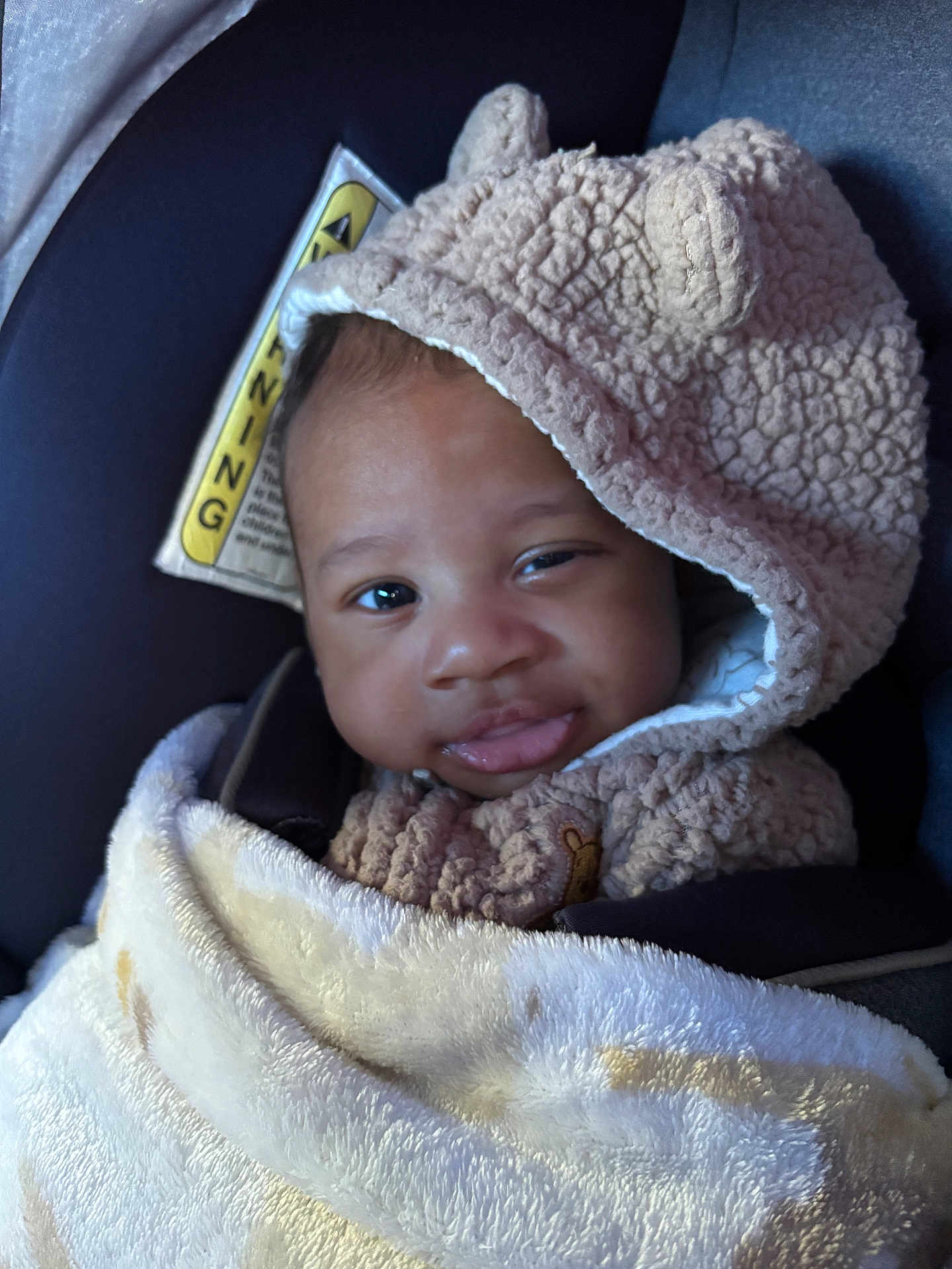 Cairo is registered to the contest to win money with this photo: baby, child, blanket, hoodie, car_seat, smile, face, cute, infant, warm_clothing, soft_texture, portrait, indoors, cozy, headwear, person, expression, seatbelt, warning_label, closeup
