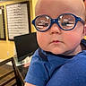 Whitley is registered to the contest to win money with this photo: baby, child, infant, glasses, eyeglasses, blue_glasses, portrait, closeup, face, serious, expression, blue_shirt, indoor, home, yellow_wall, framed_art, furniture, chair, photography, person