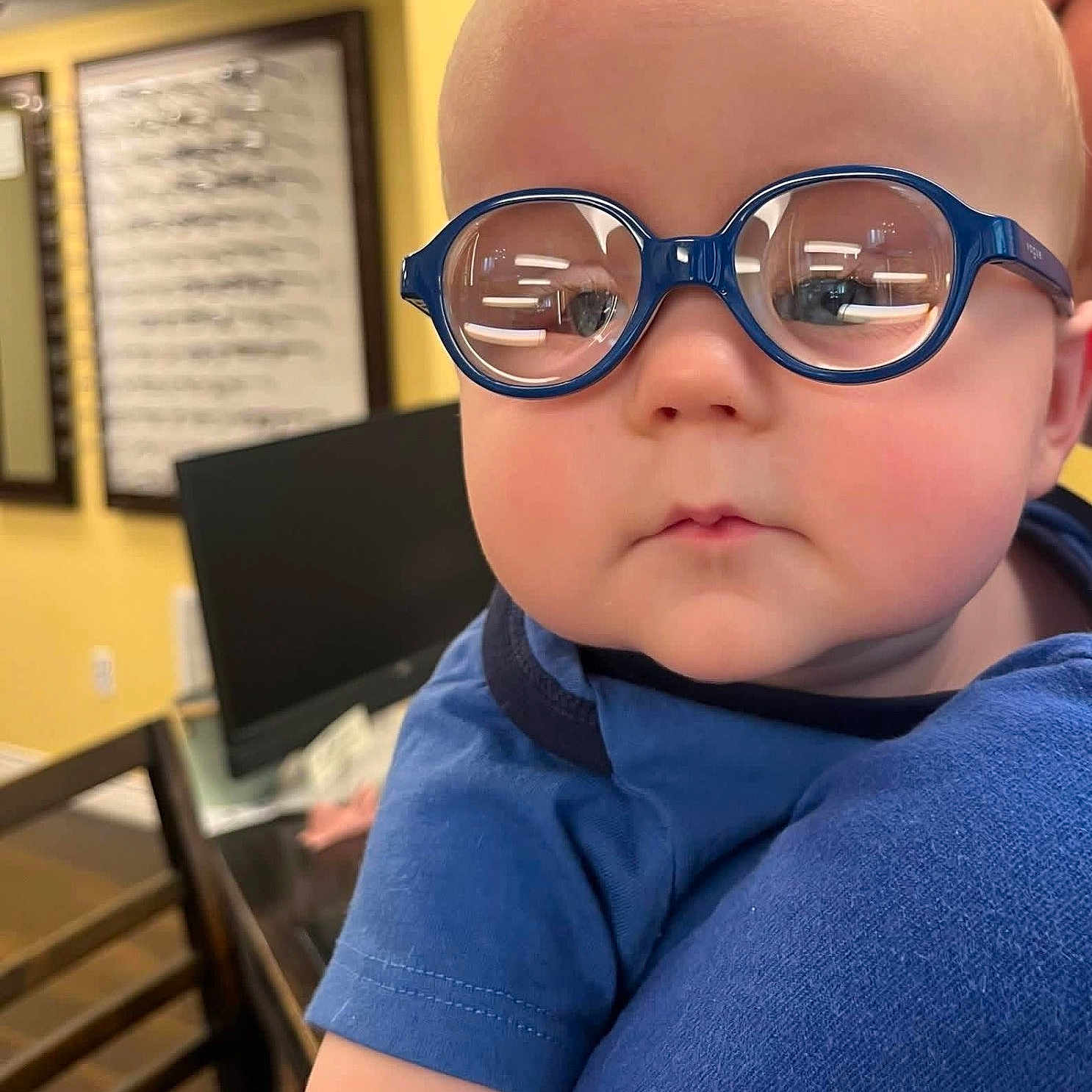 Whitley is registered to the contest to win money with this photo: baby, child, infant, glasses, eyeglasses, blue_glasses, portrait, closeup, face, serious, expression, blue_shirt, indoor, home, yellow_wall, framed_art, furniture, chair, photography, person