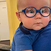 Whitley joined the competition — help win amazing prizes! baby, child, infant, glasses, blue_glasses, portrait, close_up, indoors, blue_shirt, sitting, being_held, face, head, eyes, short_hair, serious_expression, person, home, colorful_background, photography