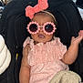 baby, child, car_seat, pink_clothing, headband, bow, sunglasses, blanket, indoor, clothing, seatbelt, hand, smiling, cute, person, infant, knitted_blanket, face, accessory, happy
