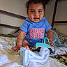 baby, child, blue_onesie, toy_truck, bed, blanket, pillow, indoor, cute, smiling, happy, curly_hair, infant, person, playing, soft_toys, cozy, home, early_childhood, portrait