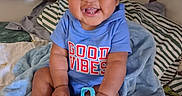 Dinero is registered to the contest to win money with this photo: baby, child, smiling, toy, blue_shirt, curly_hair, indoors, blanket, bed, happy, cute, playing, person, infant, clothing, furniture, portrait, sitting, expression, cheerful