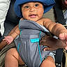 baby, blue_hat, baby_carrier, infantino, smiling, reaching_hand, adult_hand, closeup, indoor, black_leather, cushion, person, child, cute, infant, seat, support, happy, face, skin