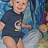 baby, bed, blue_background, child, colorful, cute, dinosaur_onesie, dolphin_mural, face, happy, indoor, infant, nursery, person, pillow, playful, smiling, toddler, toy, young_child
